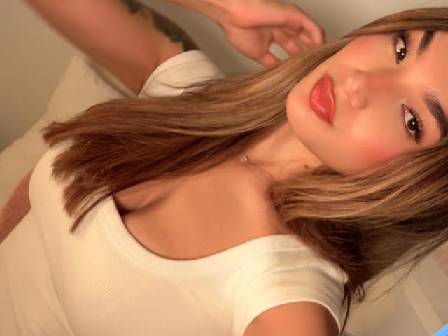 AmberMelisas has light brown hair, red lipstick, and is wearing a white tee that shows her small tits. She's looking at the camera with a slight smile.