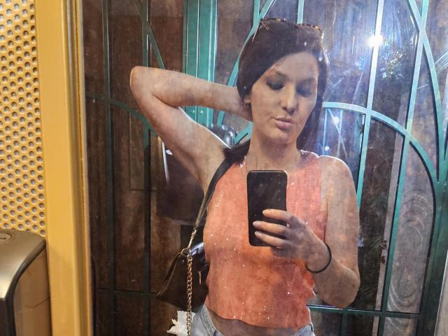 JoliDanielle with her arm raised, wearing a pink sleeveless top, holding a phone, and pouting, reflected in a dirty mirror, showing her tits and a black shoulder bag.
