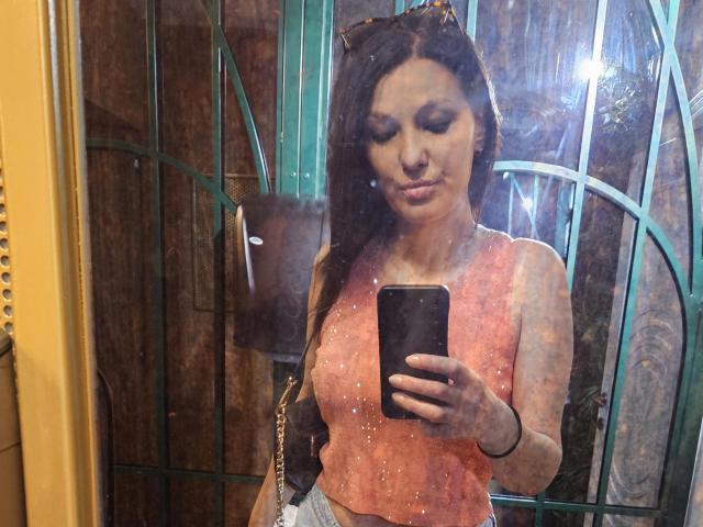JoliDanielle with long brown hair, wearing a pink sparkly top, holding a phone, reflecting in a mirror, showing her tits and ass, with green metal bars behind her.