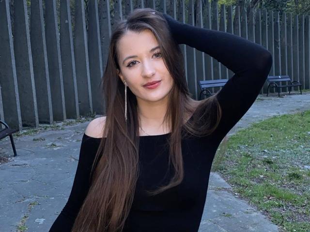 MisscharyNessy with long brown hair, black off-shoulder top, hand in hair, small earrings, slight smile, green grass, concrete path, wooden fence, black bench in background.