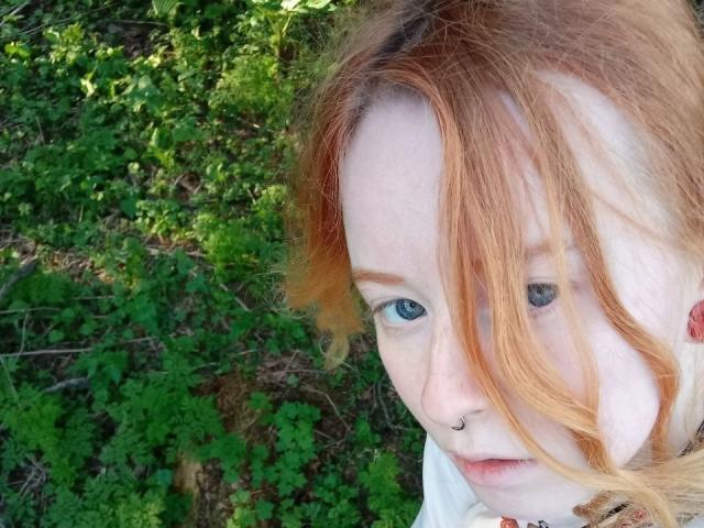 MichaelEuber with red hair, blue eyes, and a nose ring, looking sideways. Green leafy background. Wearing a white shirt. Soft light. Slightly messy hair. Pouty expression.