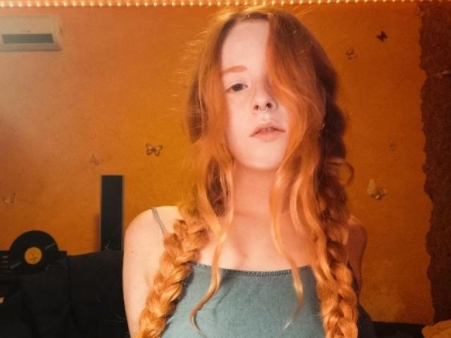 MichaelEuber with long red braids, pale skin, nose ring, wearing a blue tank top, looking slightly off-camera, in a warmly lit room. Boobs not visible.