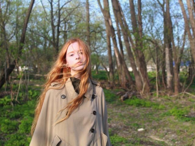MichaelEuber with long red hair, wearing a beige coat, stands in a forest. Her face shows a slightly annoyed expression. She's looking off to the side.
