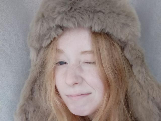 MichaelEuber has pale skin, winking with a slight smile, wearing a fluffy grey fur hat, and has dirty blonde, slightly messy hair. She's cute and has freckles.