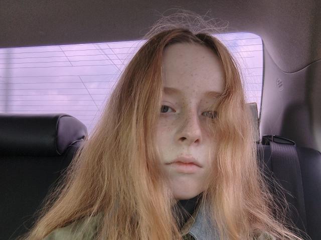 MichaelEuber has long, messy red hair, freckles, and a neutral expression, sitting in a car with black seats, wearing a green jacket. Soft light highlights his pale skin.