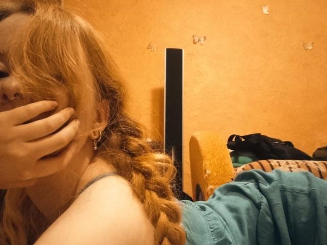 MichaelEuber with red hair, covering face with hand, showing nose ring, wearing teal shirt, in a bedroom, lying on bed with yellow blanket and black bag.