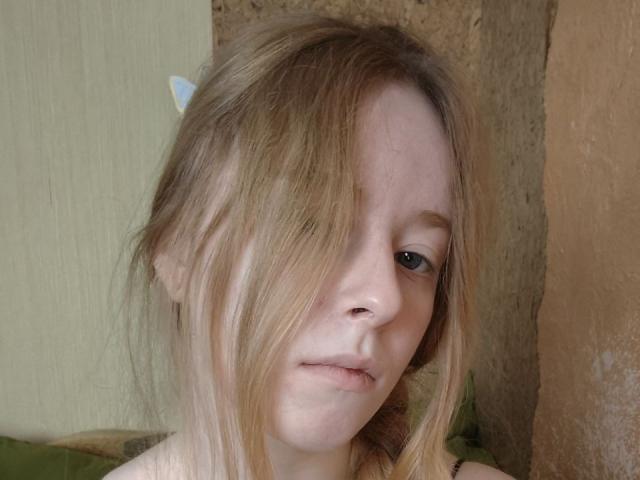 MichaelEuber's face, blonde hair covering one eye, small nose, slight smile, pale skin, green eyes, wearing a white hairclip, against a textured brown and green wall.