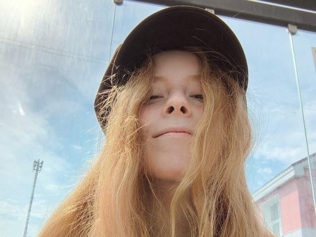 MichaelEuber with messy blonde hair, wearing a black hat, has a smug, slightly upturned nose, and a subtle smirk. Clear blue sky and glass in background.