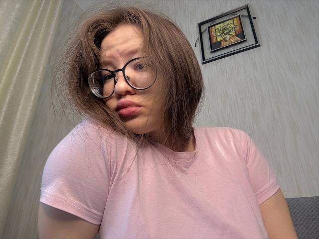 LauriceWesberry with messy brown hair, glasses, pink t-shirt, and pouty lips, looking slightly confused. Her small tits are barely visible under the shirt.