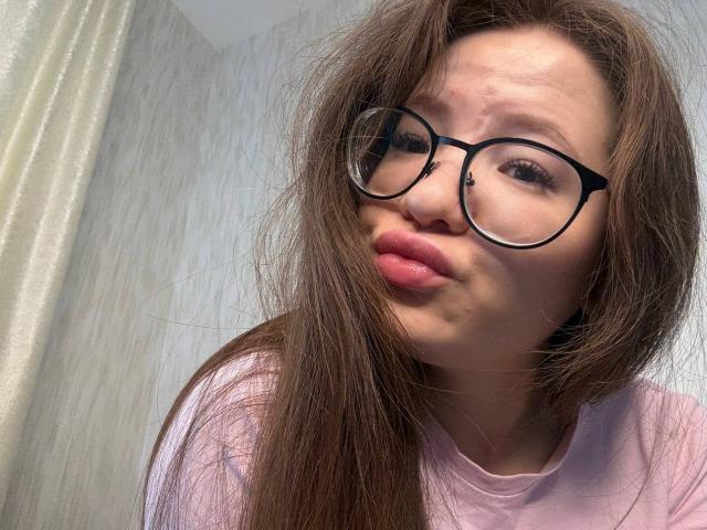 LauriceWesberry with long brown hair, wearing black glasses, pouty pink lips, and a light pink shirt, looking slightly goofy. Her ass is not visible.