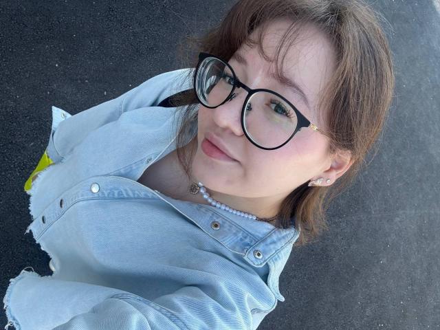 LauriceWesberry with glasses, light blue denim jacket, and pearl necklace, looking up, brown hair, subtle smile, small earrings, fair skin, slightly visible breasts, black background.