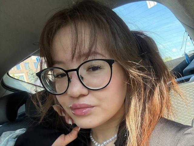 LauriceWesberry with glasses, pouty pink lips, brown hair, and a pearl necklace, sitting in a car, looking at the camera, sunlight highlighting her face.
