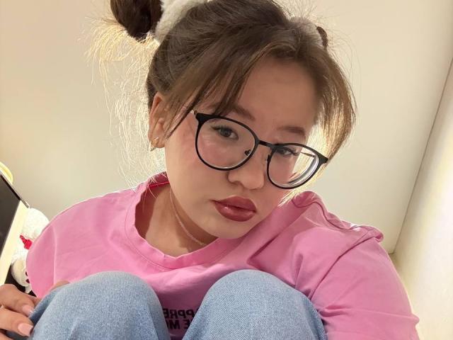 LauriceWesberry with glasses, pink shirt, and double buns, sitting with knees up, looking at camera, showing red lips, wearing blue jeans, holding a black device.