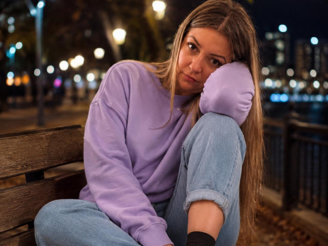 NatalyHami with long brown hair, wearing a purple sweatshirt and blue jeans, sits on a wooden bench at night, resting her head on her knee, looking pensive.