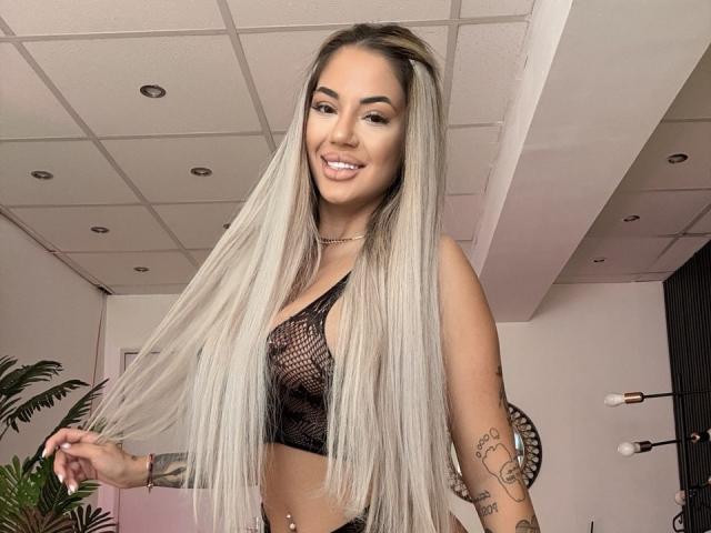 Yukenzi with long, straight, blonde hair, wearing a black mesh top, showing her small tits, smiling, tattoos on her right arm, and a belly button piercing.