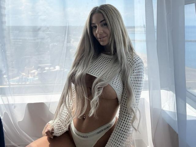 Yuukenzi with long blonde hair, wearing a white mesh top revealing her tits and white Calvin Klein panties, sitting in front of a sunlit window.