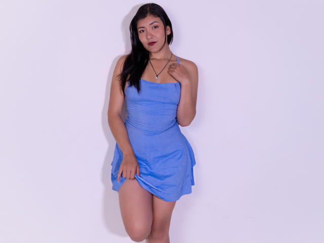 ValeryMartinez with long black hair, wearing a blue strapless dress, lifting it slightly to reveal her upper thigh, standing against a white wall, looking at the camera.