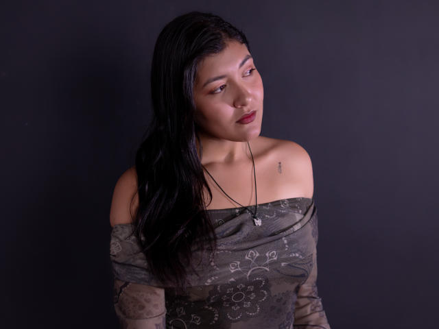 ValeryMartinez with long black hair, off-shoulder gray dress, subtle floral patterns, necklace, small tattoo on right collarbone, looking to the side, dark background.