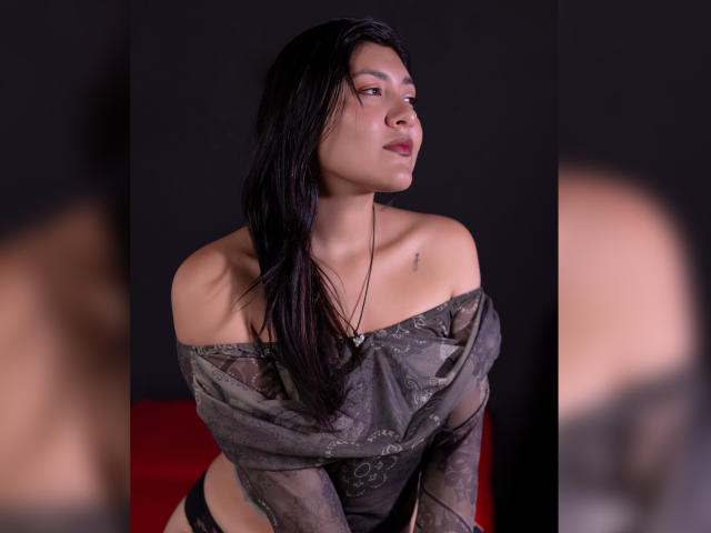 ValeryMartinez with long black hair, off-shoulder gray patterned top, small tattoo on left collarbone, looking to the side, red background, black underwear, soft lighting, slight pout.
