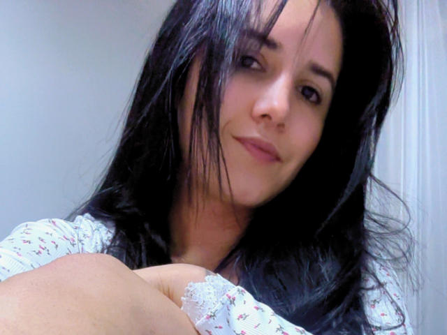 Kkatalina with black hair, light skin, and a subtle smile, wearing a floral shirt, arms crossed, showing a small lace part on her shirt.