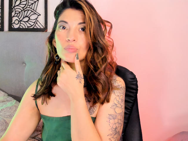 LaurentColin with wavy brown hair, green tank top, tattoos on arm, finger touching chin, black earrings, sitting in black chair, pink and green lighting, pouty expression.