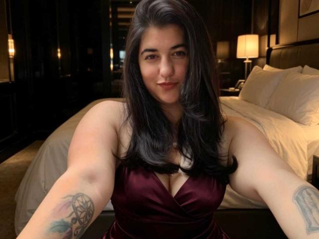 Danny Waller, con lunghi capelli neri, indossa un abito color borgogna scuro, mostrando tatuaggi su entrambe le braccia, sorride e sta scattando un selfie in una camera da letto poco illuminata, con un letto sullo sfondo..
