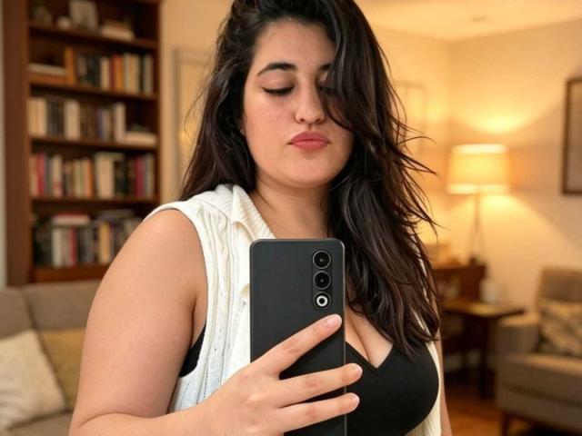 Danny Waller, con capelli scuri e ondulati, indossa una canotta bianca e una maglietta nera, tiene in mano un telefono nero, mostra il seno, in una stanza con una luce soffusa..