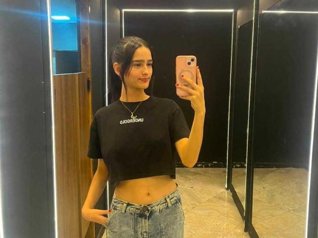 MelodyMansson with dark hair, black crop top, showing her midriff, and blue jeans, taking a mirror selfie, holding a pink phone, wearing a necklace, smiling slightly.