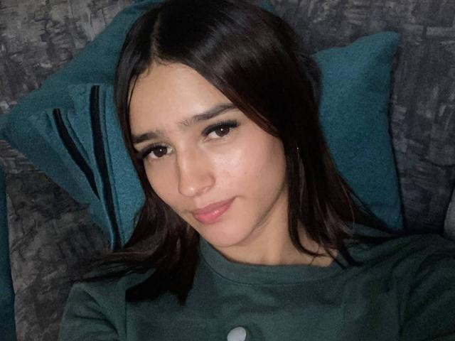 MelodyMansson has dark brown hair, light skin, and brown eyes. She's wearing a green shirt, looking slightly to the side with a subtle smile, and has pink lipstick.