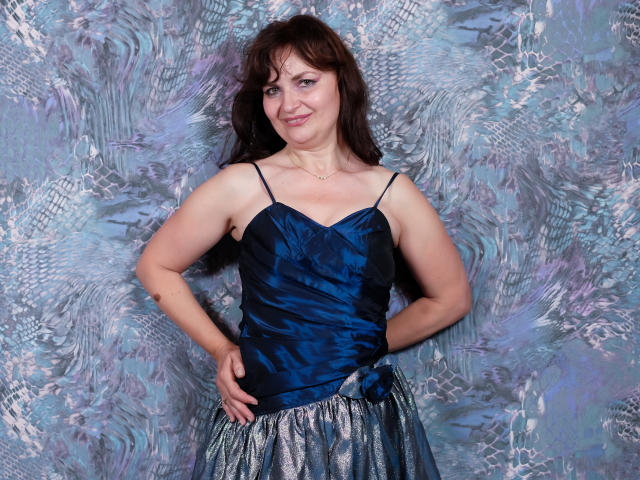 AngieGreen with medium tits, wearing a blue satin top and silver shimmer skirt, hands on hips, smiling, brown hair, standing against a textured blue-gray wall.
