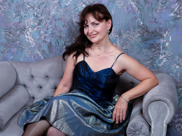 AngieGreen with medium tits, wearing a blue satin dress and green earrings, sits on a gray tufted sofa, smiling at the camera, with a shiny blue skirt.