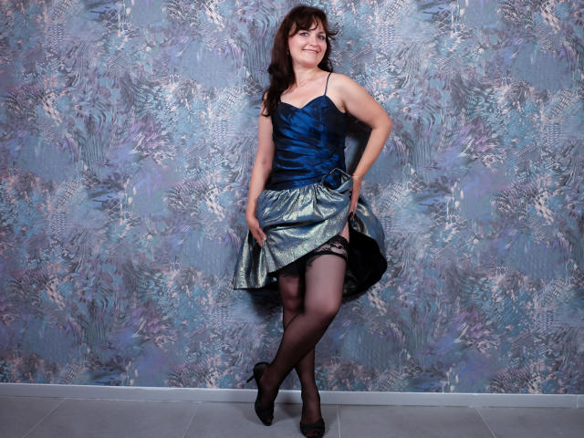 AngieGreen in a blue satin top and shiny green skirt, showing black lace stockings and black heels, standing with a smile, hands on skirt, her ass slightly raised.
