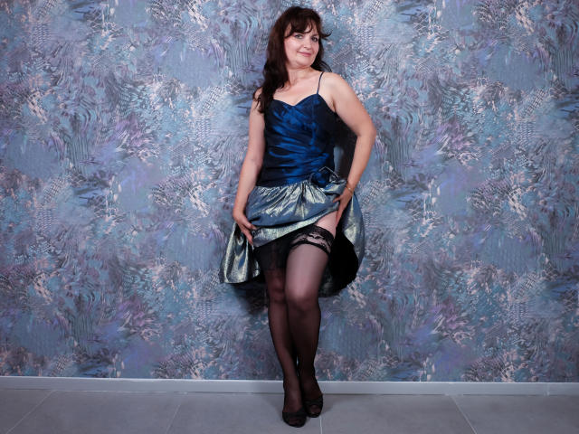 AngieGreen with dark brown hair, wearing a blue dress, lifting it to reveal black lace stockings and a garter, standing in black heels, smiling at the camera.
