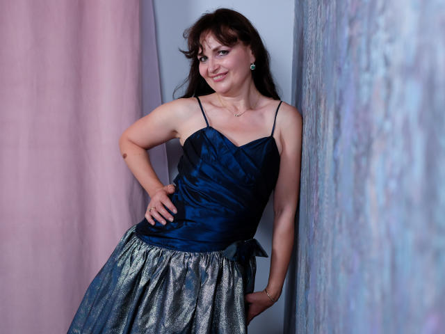 AngieGreen with medium tits, wearing a blue satin top and silver sparkly skirt, standing with hand on hip, smiling, brown hair, blue earrings, pink curtain on left.