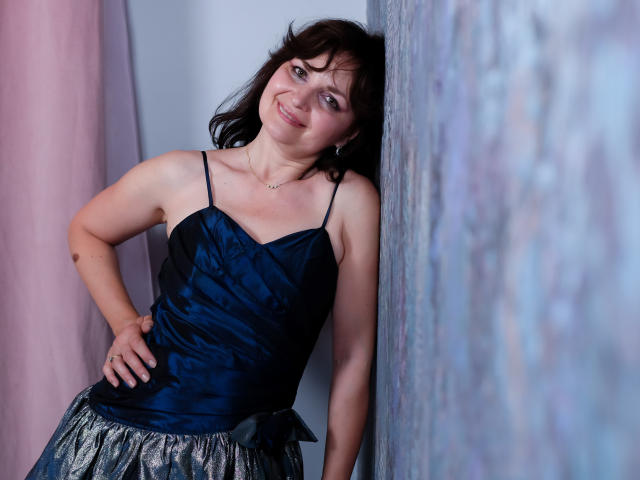 AngieGreen with dark brown hair, smiling, wearing a navy blue satin top and silver sequined skirt, leaning against a textured wall, hand on hip, showing her small tits.