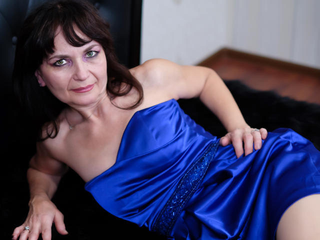 AngieGreen with green eyes, brown hair, and pink lipstick, wearing a shiny blue dress with a black lace belt, lying on a black furry surface, showing her tits.