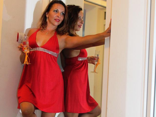 Aprel with medium tits, red dress, and a silver belt, holding a glass of champagne, leaning against a mirror, smiling, her reflection shows her sexy pose.