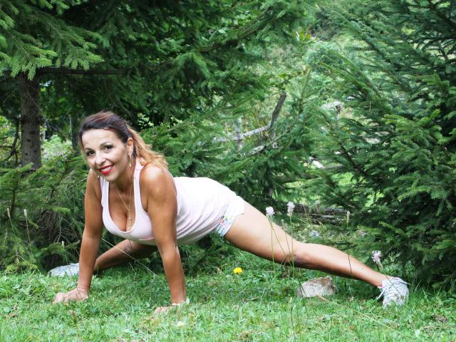 Aprel is on all fours, doing a plank in a forest, wearing a pink tank top and colorful shorts, with red lipstick and a smile, showing her tits and ass.