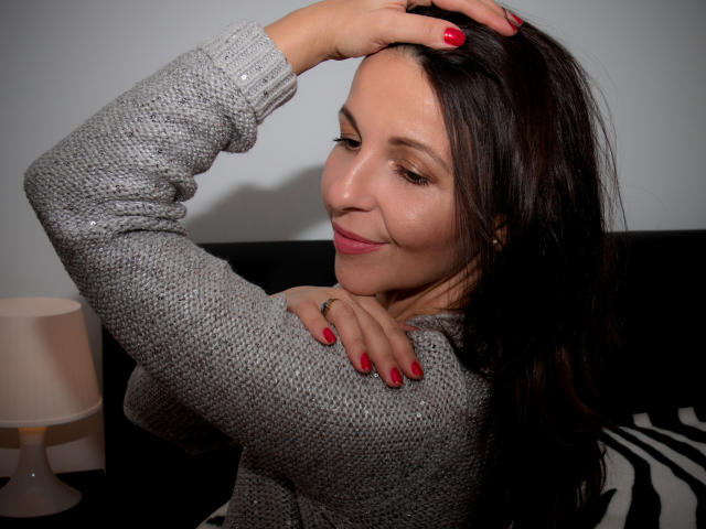 Aprel with dark hair, red nails, and a gray knit sweater, smirking while hand in hair, exposing small tits and cute face. Soft lighting, zebra-patterned pillow.