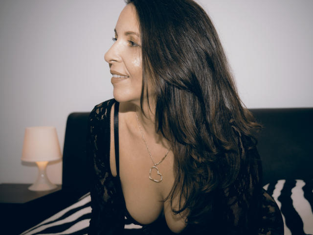 Aprel with long brown hair, wearing a black lace top, showing her cleavage, smiling, looking to the side, wearing a heart necklace, by a lamp on a zebra-pattern bed.