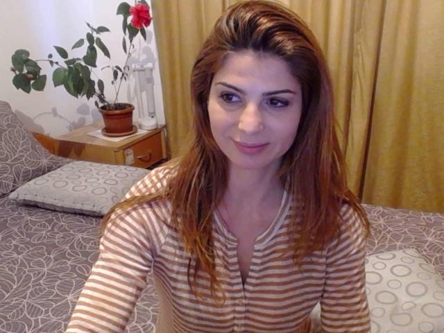 Sexydollhotx with brown hair, striped shirt, sitting on bed, looking at camera, smiling, small pink nipples visible, yellow curtains in background, potted plant on wooden nightstand.