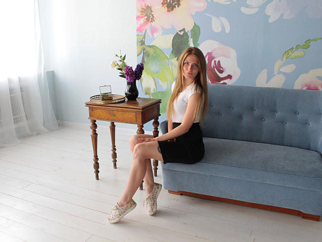 GretyArdent sits on a blue sofa, wearing a white top, black skirt, and white shoes, with legs crossed, displaying her ass, and a vase with purple flowers on a table.