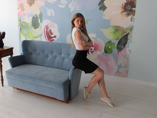 GretyArdent with small tits, sitting on blue couch, wearing white crop top, black skirt, and white sneakers, arms crossed, in room with floral wallpaper, white floor.