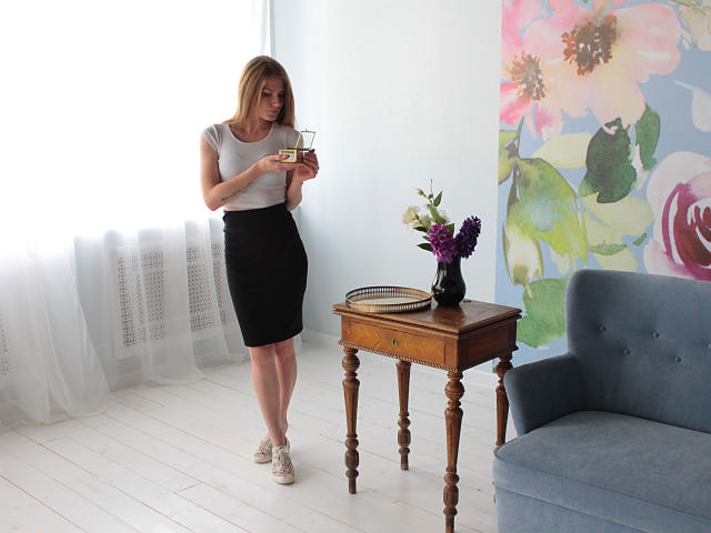 GretyArdent with blonde hair, gray top, black skirt, and white sneakers, holding a cup, standing by a wooden table with flowers and a book, next to a blue chair.