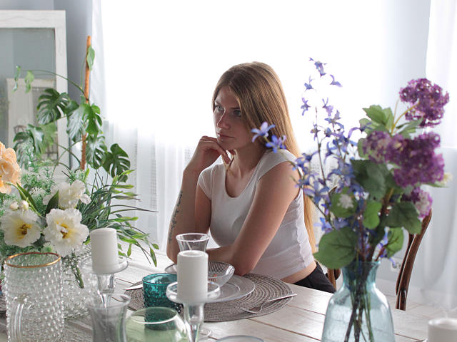 GretyArdent with long brown hair, white t-shirt, and glasses, sitting at a table with flowers, candles, and green glass jar, looking pensive. Her tits are small, arms tattooed.