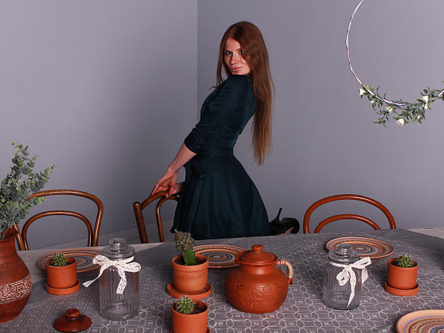 GretyArdent stands with long brown hair, wearing a dark green dress, looking over her shoulder. Table with terracotta pots, glass jars, and patterned gray cloth. Ass, tits, potted plants.