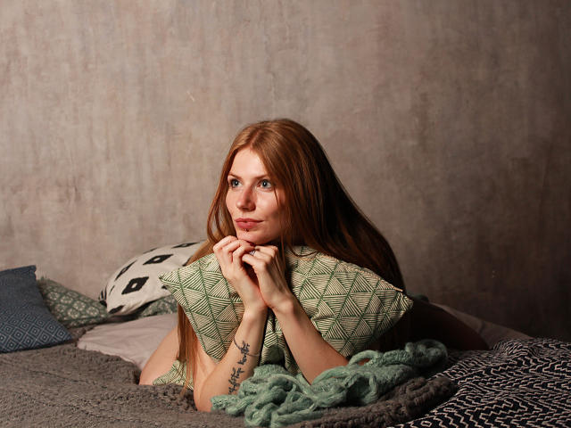 GretyArdent with long red hair, holding a green patterned pillow, looking at the camera with a slight smile, lying on a bed with gray and green blankets.