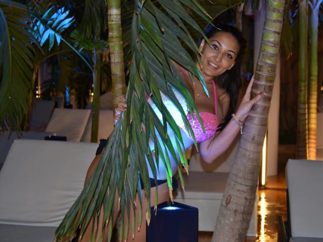 AlesyaNoova with medium tits, wearing a pink lace top, black shorts, and sunglasses, hiding behind a palm tree, smiling, holding the tree with her right hand.