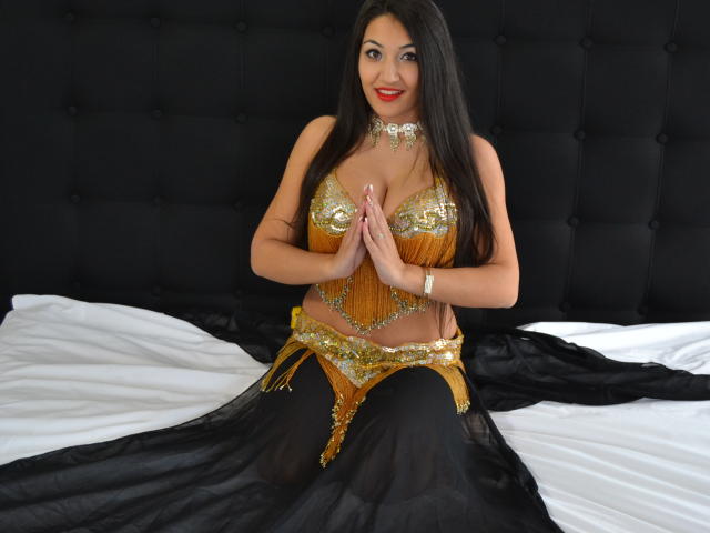 AlesyaNoova with long black hair, red lipstick, gold belly dancer outfit, and black harem pants, sitting on a bed, hands together, smiling, showing her shiny tits and belly.