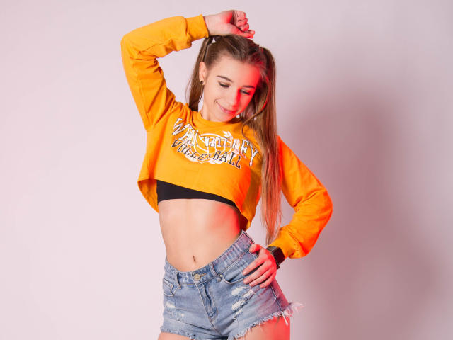 LucyBestCandy with long blonde hair, wearing an orange crop top, black bra, and ripped denim shorts, smiling, hand in hair, showing her toned midriff and cute ass.
